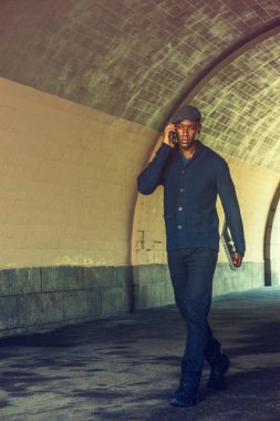 Genç Afrikalı-Amerikalı adam giyiyor mavi şal yaka hırka kazak, siyah pantolon, spor ayakkabı, dizüstü bilgisayar holding, cep telefonuyla konuşurken, yeni Yor sokak Köprüsü altında yürüyüş düz kapak