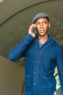 Genç yakışıklı Afrikalı-Amerikalı adam, New York şehrinde seyahat mavi kitted şal yaka hırka kazak giyen gri düz kapak sokak Köprüsü'nün altında yürüyüş cep telefonuyla konuşurken, desenli