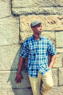 Genç yakışıklı Afrikalı-Amerikalı adam New York'ta giyiyor mavi ve beyaz desenli gömlek, bej pantolon, gri kaya duvarı güçlü güneş altında karşı duran, uzağa arıyorsunuz, düşünme düz cape, desenli