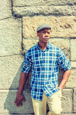 Genç yakışıklı Afrikalı-Amerikalı adam New York'ta giyiyor mavi ve beyaz desenli gömlek, bej pantolon, gri kaya duvarı altında güçlü güneş, düşünme, lost ın düşünce karşı ayakta düz cape desenli