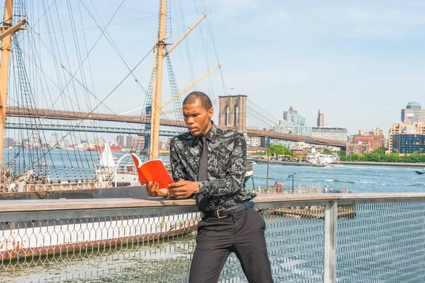African American businessman traveling in New York. Wearing black flower patterned shirt, tie, a young guy standing at harbor, reading red book. Manhattan, Brooklyn bridges, boats on background