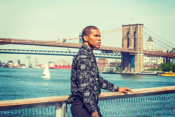 African American businessman traveling in New York. Wearing black flower patterned shirt, tie, a young guy standing at harbor, thinking. Manhattan, Brooklyn bridges on background