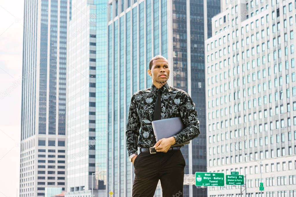 African American businessman working in New York. Wearing black flower patterned shirt, tie, holding laptop computer, young  guy standing in front of business district, confidently looking forward