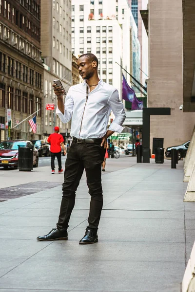 Young African American Man with beard, wearing white shirt, black pants, leather shoes, standing on street in Manhattan, New York, reading messages on cell phone. People, cars, buildings on backgroun