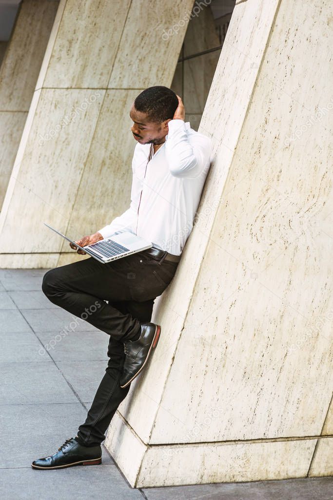 Young African American businessman working in New York, wearing white shirt, black pants, leather shoes, standing against column outside, working on laptop computer, scratching head, reading, thinking