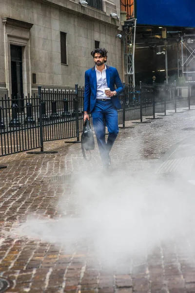 Raining day - grainy, foggy, wet feel. Young East Indian American Business Man with beard, wearing blue suit, white shirt, carrying leather hand bag, holding coffee cup, walking on street in New York