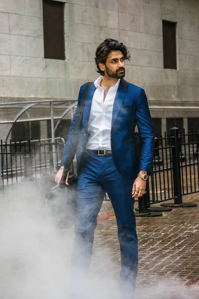 Raining day - grainy, foggy, wet feel. Young East Indian American Business Man with beard, wearing blue suit, white shirt, carrying leather shoulder bag, looking away, walking on street in New York