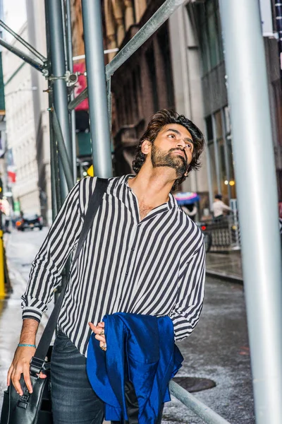 Raining day - grainy, drizzling, wet feel. Young East Indian American Man with beard, wearing black, white striped shirt, carrying leather shoulder bag, carrying blue clothes, looking up in New York