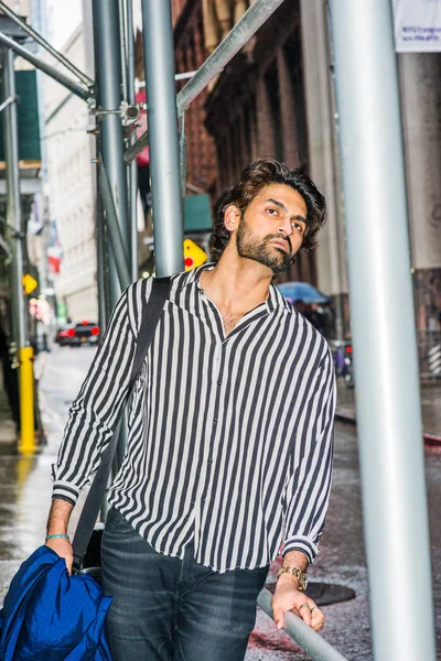 Raining day - grainy, drizzling, wet feel. Young East Indian American Man with beard, wearing black, white striped shirt, carrying shoulder bag, holding blue clothes, standing on street in New York