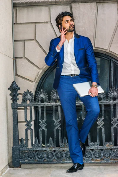 Sakallı, mavi ceketli, beyaz gömlekli, mavi pantolonlu, siyah deri ayakkabılı, dizüstü bilgisayarlı, New York 'taki ofisin önünde duran, cep telefonuyla konuşan genç bir işadamı.