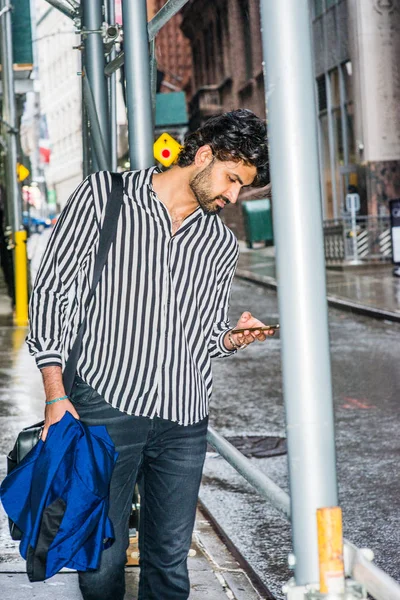 Raining day - grainy, drizzling, wet feel. Young East Indian American Man with beard traveling in New York, wearing black, white striped long sleeve shirt, standing on street, looking down, texting