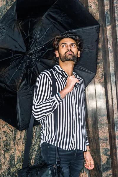 Raining day - grainy, drizzling, wet feel. Young East Indian American Man with beard, wearing black, white striped shirt, carrying bag, holding umbrella, standing outside in New York, looking up