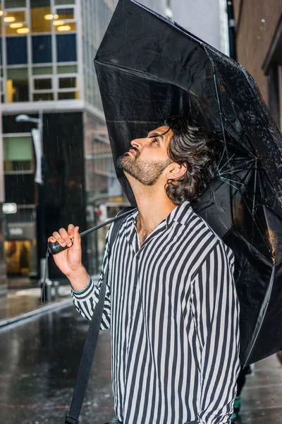 Yağmur günü - grenli, yağmurlu, ıslak hissediyorum. Sakallı genç Doğu Hint Amerikalı Adam, siyah, beyaz çizgili gömlek giyen, şemsiye taşıyan, sokakta duran, başını kaldıran, yukarı bakan