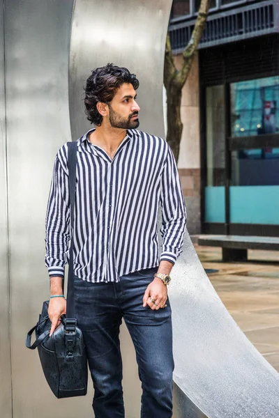 Raining day - grainy, drizzling, wet feel. Young East Indian American Man with beard, wearing black, white striped shirt, black pants, carrying shoulder bag, standing on street in New York, looking