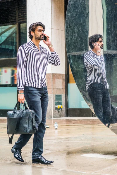 Raining day - grainy, drizzling, wet feel. Young East Indian American Man with beard, wearing striped shirt, black pants, leather shoes, holding bag, walking on street by mirror, talking cell phone