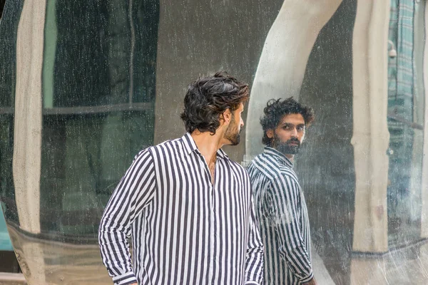 Raining day - grainy, drizzling, wet feel. Young East Indian American Man in New York, with beard, wearing black, white striped shirt, standing outside, turning around, looking reflection on mirror