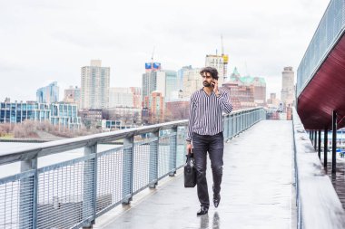 Yağmur - grenli, yağmurlu, ıslak hissediyorum. Çizgili gömlek giyen, siyah pantolon, deri ayakkabı giyen, el çantası tutan, New York'ta seyahat eden, cep telefonuyla konuşan genç işadamı. Brooklyn uzak arka planda