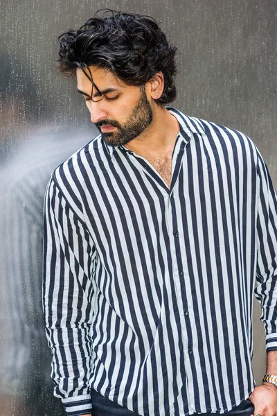 Raining day - grainy, drizzling, wet feel. Young East Indian American Man in New York, with beard, wearing black, white striped shirt, standing outside against silver metal wall, looking down, thinks