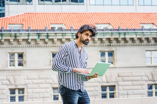 Raining - grainy, drizzling, wet feel. Young East Indian American Businessman with beard, wearing black, white striped shirt, standing outside office building in New York, working on laptop computer