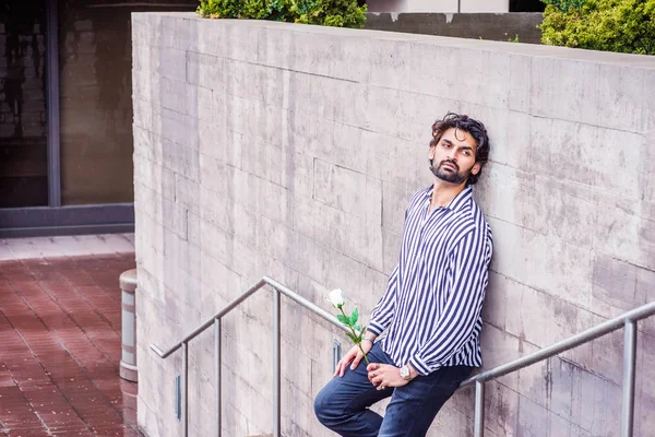 Raining day - grainy, drizzling, wet feel. Young East Indian American Man with beard, wearing black, white striped shirt, holding white rose, standing against wall in New York, looking away, thinking