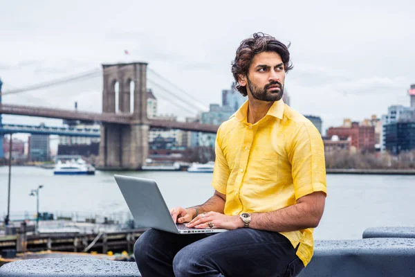 Raining - grainy, drizzling, wet feel. Young Man with beard traveling in New York, wearing short sleeve yellow shirt, sitting by East River, working on laptop computer. Bridges, boat on background