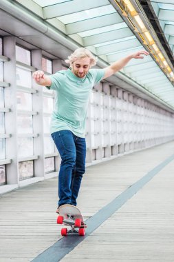 New York'ta okulda paten kaymak. Uzun sarı saçlı genç üniversite öğrencisi, açık yeşil t shirt giyiyor, mavi kot pantolon, kapalı geçit ahşap zemin üzerinde kaykay üzerinde duran, denge tutmak, paten