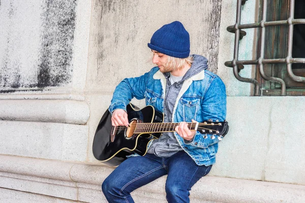 Street Musician in New York City. Young college student with long blonde hair, wearing blue Denim jacket coat, knitted hat, sitting against wall by window on campus, looking down, playing guitar