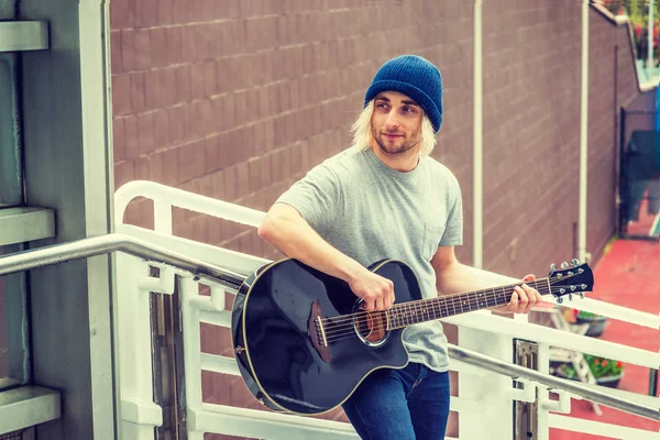 Young college student playing music in New York City. Young Man with long blonde hair, wearing knitted hat, gray T shirt, standing against railing outside on campus , playing guitar