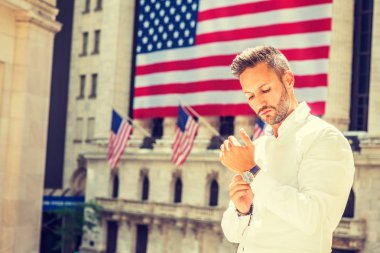 Young European Man with beard traveling in New York in summer, wearing long sleeve white shirt, wristwatch, standing office building with American flags under sun, looking down, hand buttoning cuff