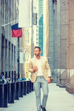 Young handsome European Man with beard traveling in New York City. Young guy wearing beige blaze, white shirt, gray pant, walking on narrow vintage street with banners, looking around, thinking