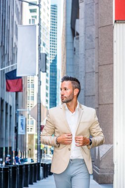 Young handsome European Man with beard traveling in New York City. Young guy wearing beige blaze, white shirt, gray pant, walking on narrow vintage street with banners, looking around, thinking.