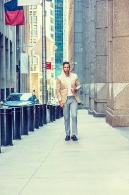 Young handsome European Man with beard traveling in New York City. Young guy wearing beige blaze, white shirt, gray pant, black leather shoes, walking on narrow vintage street with banners, cars.