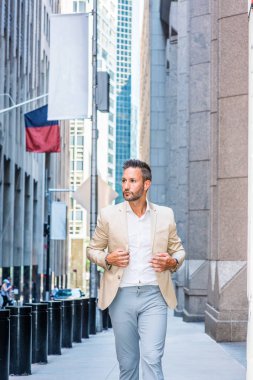 Young handsome European Man with beard traveling in New York City. Young guy wearing beige blaze, white shirt, gray pant, walking on narrow vintage street with banners, looking around, thinking.