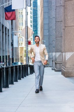 Young handsome European Man with beard traveling in New York City. Young guy wearing beige blaze, white shirt, gray pant, black leather shoes, walking on narrow vintage street with banners, cars