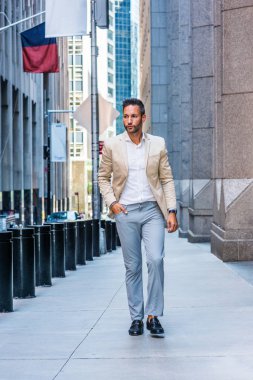 Young handsome European Man with beard traveling in New York City. Young guy wearing beige blaze, white shirt, gray pant, black leather shoes, walking on narrow vintage street with banners, cars.