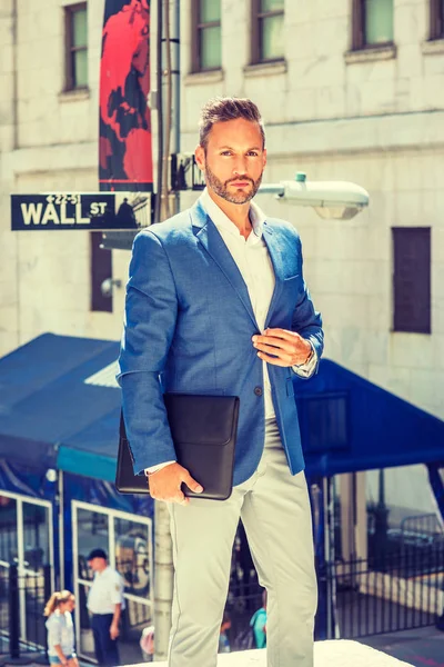 Young European Businessman with beard traveling, working in New York City, wearing blue blazer, white shirt, holding briefcase, standing on street outside office by Wall Street sign, looking forward