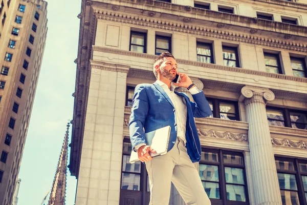 Sakallı genç Avrupalı iş adamı, New York 'ta çalışıyor, mavi ceket giyiyor, gri pantolon giyiyor, dizüstü bilgisayarı tutuyor, eski ofis binasının önünde duruyor, cep telefonunu arıyor.