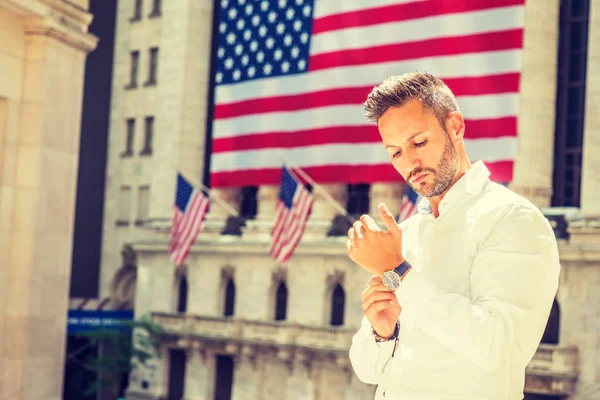 Young European Man with beard traveling in New York in summer, wearing long sleeve white shirt, wristwatch, standing office building with American flags under sun, looking down, hand buttoning cuff