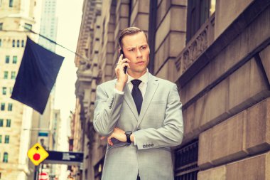 New York 'ta seyahat eden genç bir iş adamı gri ceket, beyaz gömlek, siyah kravat, kol saati, eski caddede yüksek binalarla yürüyor, bayrak çekiyor, cep telefonuyla konuşuyor, kaşlarını çatıyor, düşünüyor.