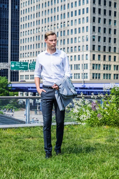 Young Handsome European Man traveling in New York City, wearing white shirt, black pants, leather shoes, arm carrying jacket, standing on green lawn in business district with high modern buildings