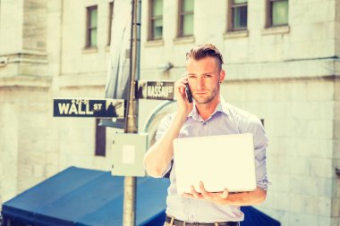 Genç yakışıklı adam seyahat, New York'ta çalışan, uzun kollu giyiyor, açık renkli gömlek, güneş altında Wall Street işareti ofis dışında duran, dizüstü bilgisayar üzerinde çalışan, cep telefonu konuşuyor