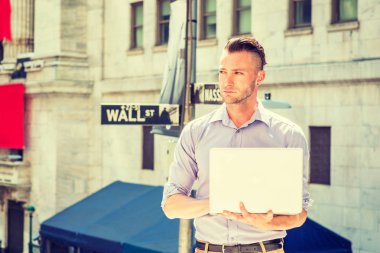 Genç yakışıklı adam seyahat, New York'ta çalışan, açık renk giyiyor, uzun kollu gömlek, Wall Street işareti ofis dışında duran, güneş altında, dizüstü bilgisayar üzerinde çalışan, seyir, düşünme