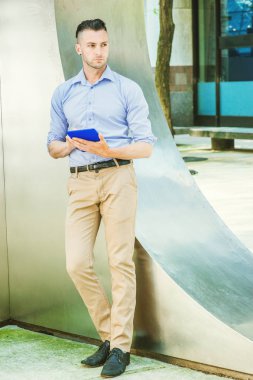 New York'ta dışarıda kitap okuyan, açık mor, uzun kollu gömlek, bej pantolon, siyah ayakkabı giyen, sokakta gümüş metal duvar da duran, elleri mavi tablet bilgisayar tutan, düşünme