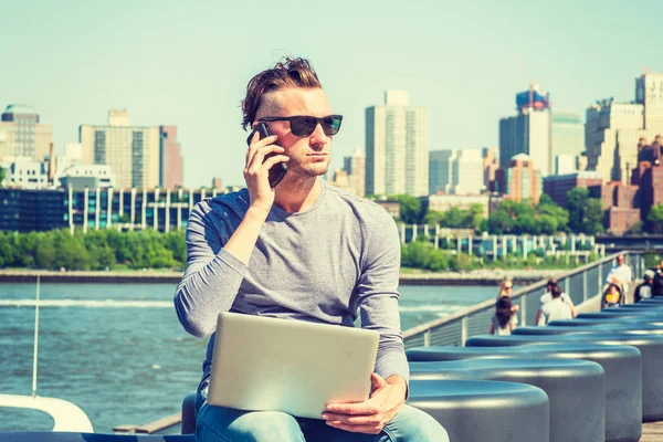 New York'ta çalışan, gri, uzun kollu tişört giyen, güneş gözlüğü takan, nehir kıyısında oturan, dizüstü bilgisayarda çalışan, cep telefonuyla konuşan amerikalı genç bir adam. Arka planda Brooklyn binaları