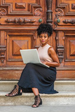 Afro-Amerikan Öğrenci New York'ta eğitim, afro saç modeli ile, kolsuz açık renk üst giyiyor, siyah skeç, askılı sandalet, New York'ta ofis kapısı başında oturan, dizüstü bilgisayar hesaplama üzerinde çalışan