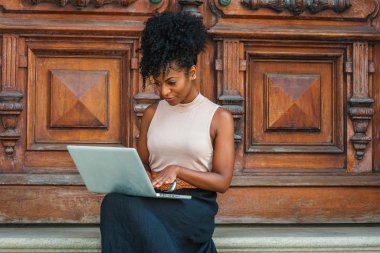 Başarıya Giden Yol. Afro saç modeli ile genç Afrikalı Amerikalı kadın kolsuz açık renk üst giyen, siyah skeç, New York'ta vintage ofis kapısı tarafından oturan, aşağı bakıyor, dizüstü bilgisayar üzerinde çalışıyor