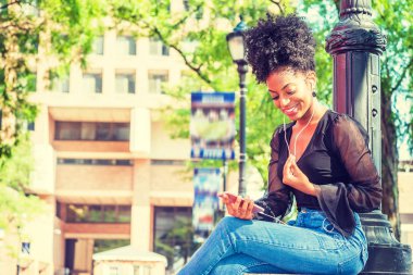 Afro saç modeli ile Genç Afro-Amerikan Kadın örgü sırf uzun kollu gömlek bluz giyen, mavi kot pantolon, New York'ta kampüste ışık direği tarafından oturan, kulaklık ve cep telefonu ile müzik dinleme