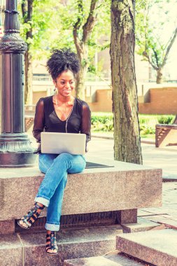 Afro saç modeli ile Genç Afro-Amerikan Kadın örgü sırf uzun kollu gömlek giyiyor, kot pantolon, New York kampüsünde ışık direği tarafından oturan, kulaklık ile müzik dinleme, dizüstü bilgisayar üzerinde çalışan.