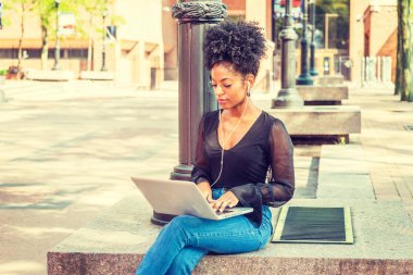 Afro saç modeli ile Genç Afro-Amerikan Kadın örgü sırf uzun kollu gömlek giyiyor, kot pantolon, New York kampüsünde ışık direği tarafından oturan, kulaklık ile müzik dinleme, dizüstü bilgisayar üzerinde çalışan