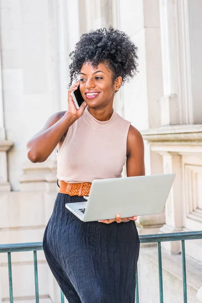 African American Businesswoman working in New York. Young black college student with afro hairstyle sitting on railing in vintage style office building, working on laptop computer, making phone call.